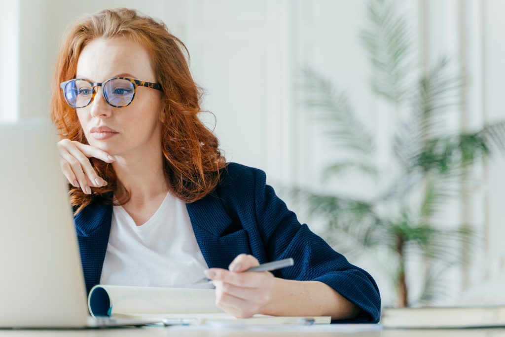 Serious experienced female coach makes records in notepad, focused at screen of laptop